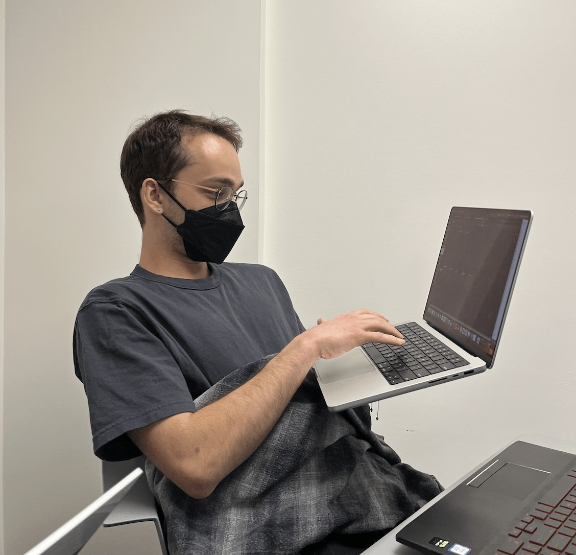 A Caucasian man in a black mask and circlular rimmed glasses sits in a chair while holding a laptop up with his left hand and tapping on the keyboard with his right.