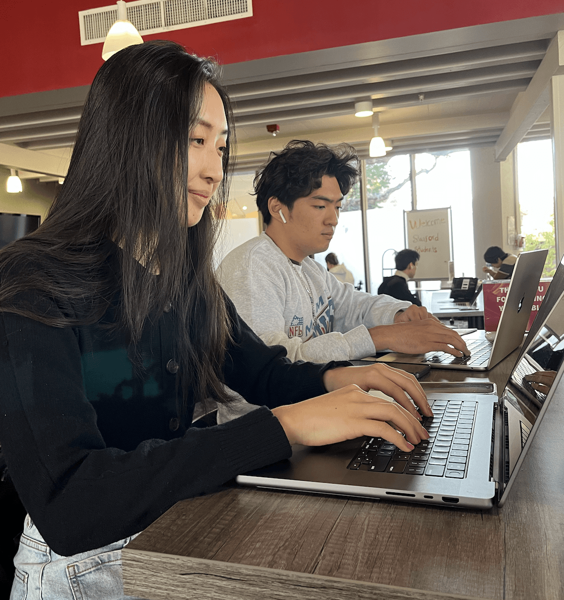 A young Asian woman types on a laptop with a small smile on her face. A young Asian man next to her with earbuds in does the same.