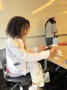 A woman with curly dark hair sits at a desk smiling. She looks at a white index card she's holding. A few other index cards sit on the table in front of her.