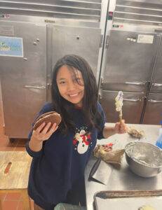 Candy is wearing a blue sweater and standing in the kitchen next to the counter. She is smiling while holding a spatula in one hand and a large, red velvet sandwich cookie in the other hand. 
