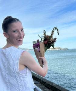 Woman holding flowers near water, smiling at camera.